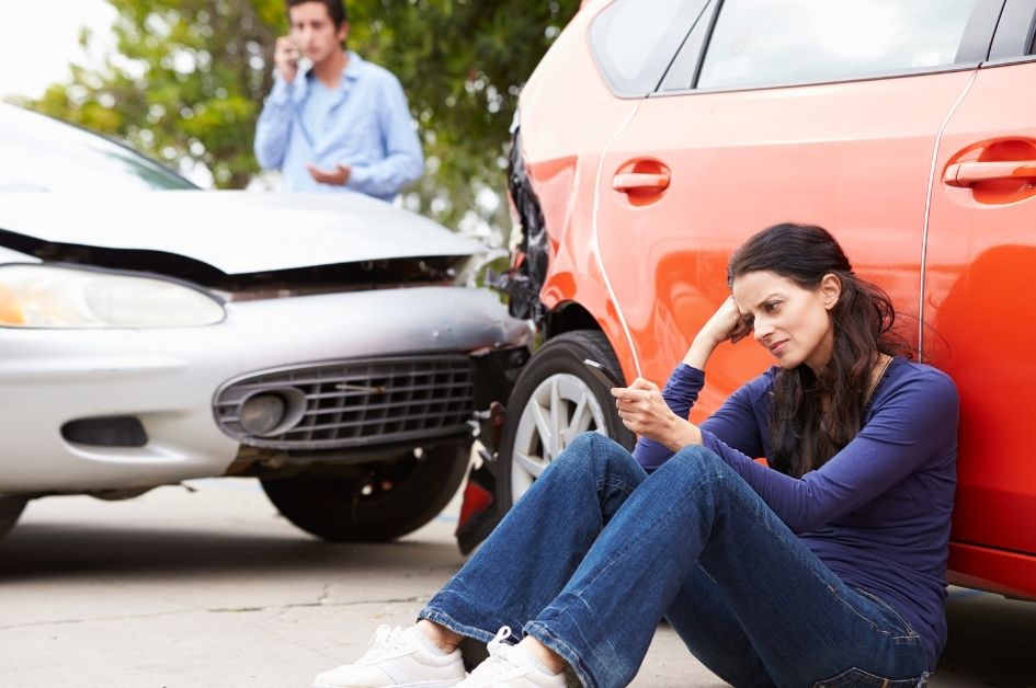 2 peple in a car collision, one woman leaning against an orange car; one man in the background on the phone
