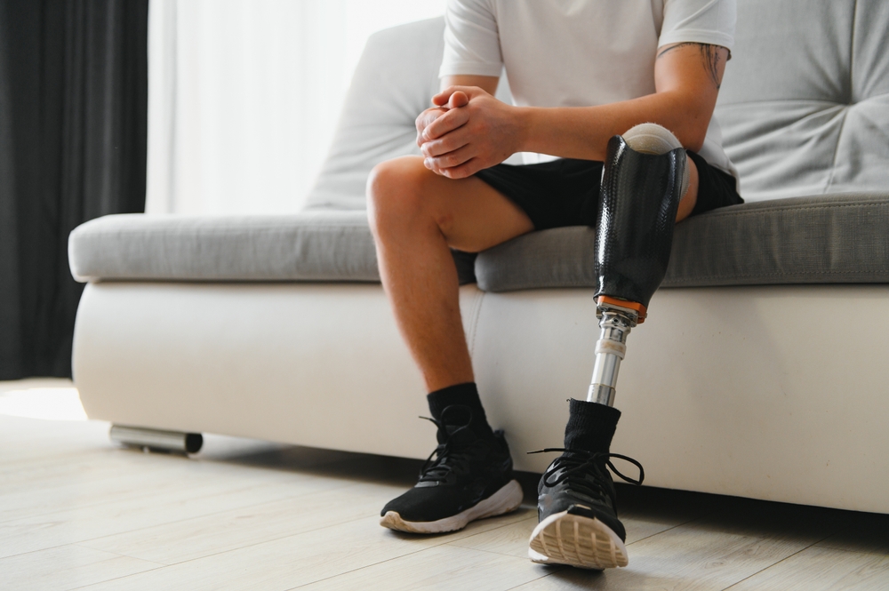 Man amputee with prosthetic leg disability on above knee transfemoral leg prosthesis wearing shoe sitting on sofa, close up. People with amputation disabilities everyday life concept. (2)
